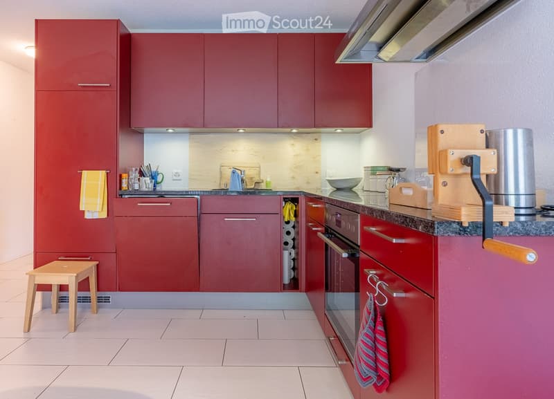 Modern kitchen with red cabinets, white tiled floor, and stainless steel appliances. Features include a small wooden stool, a coffee maker on the counter, and a hanging towel.