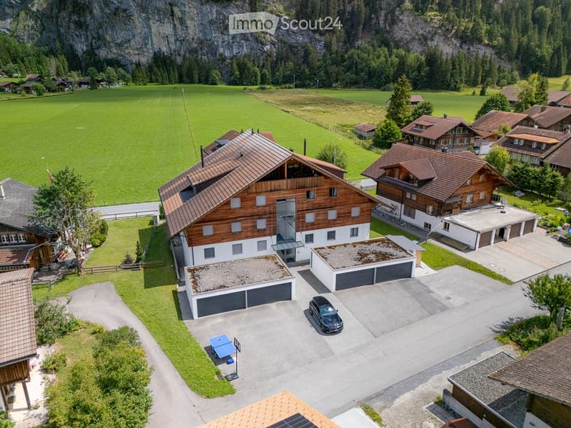 Aerial view of a large residential complex with multiple houses, garages, and a parking area, surrounded by green fields and mountains.