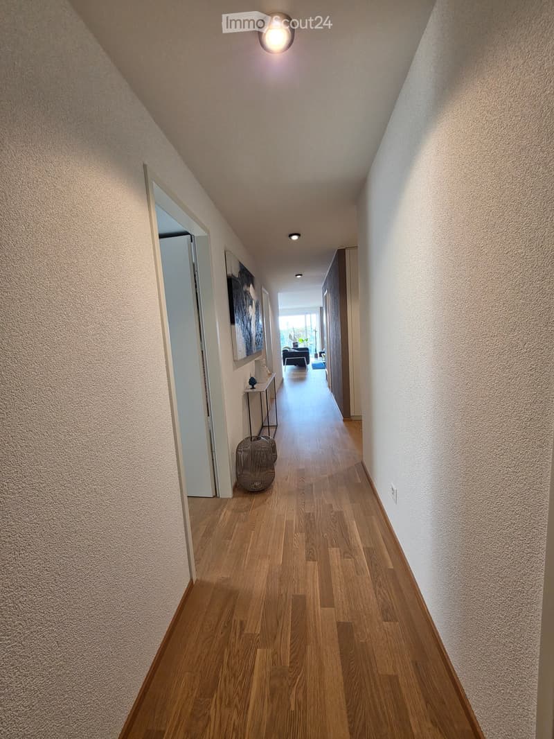 A modern hallway with wooden flooring, white walls, and recessed lighting. Features a small table with a decorative item and a woven basket on the floor.