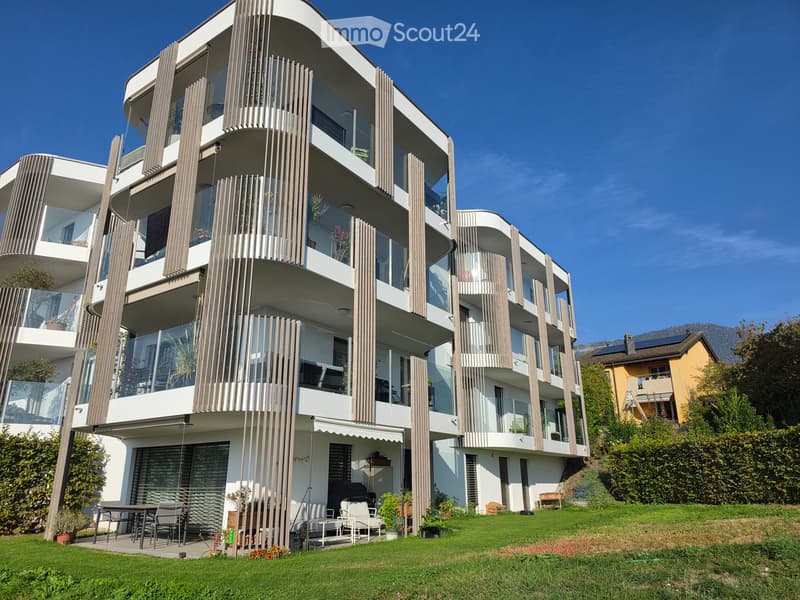 Modern multi-story building with balconies, large windows, and a raised ground floor, surrounded by a well-maintained lawn and outdoor seating area.