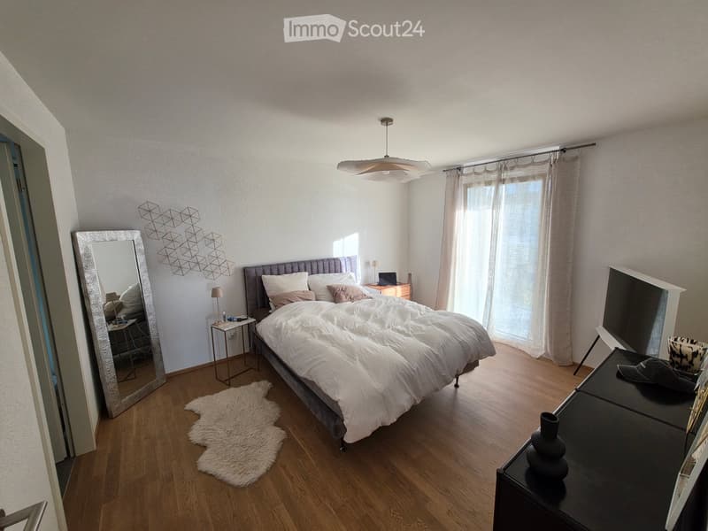 Modern bedroom with a large bed, white bedding, a fluffy white rug, a large mirror, a nightstand with a lamp, and a small table with a laptop and decorative items.