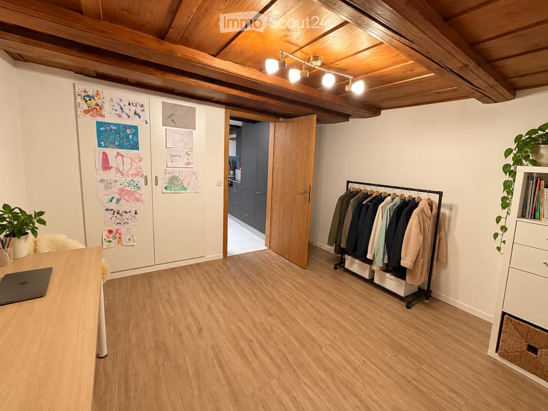 A well-lit room with wooden flooring and exposed wooden beams on the ceiling. Features include a laptop on a table, a coat rack with jackets, a bookshelf with books and plants, and several colorful children's drawings on the wall.