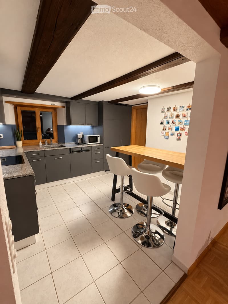 Modern kitchen with grey cabinets, white tiled floor, and a wooden dining table with white stools. Features a sink, microwave, coffee maker, and a potted plant on the counter.