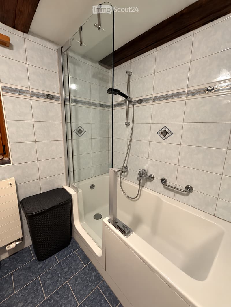 Modern bathroom with a white bathtub, glass-enclosed shower, and a wicker basket on the floor. The walls are tiled with light-colored tiles and decorative accents.