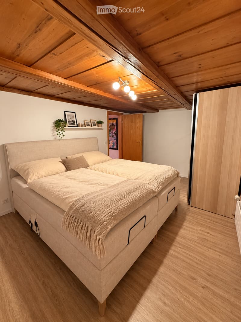 Cozy attic bedroom with wooden beams, a large bed with pillows and a blanket, a shelf with picture frames and a small plant, and warm lighting.