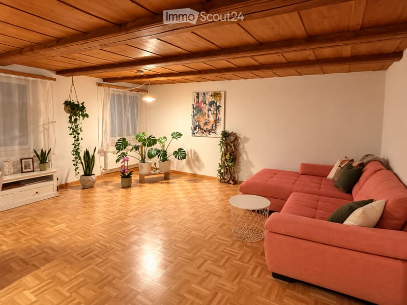 Living room with wooden herringbone flooring, exposed wooden ceiling beams, large window with sheer curtains, red sectional sofa, small round white coffee table, several potted plants, and a decorative picture on the wall.