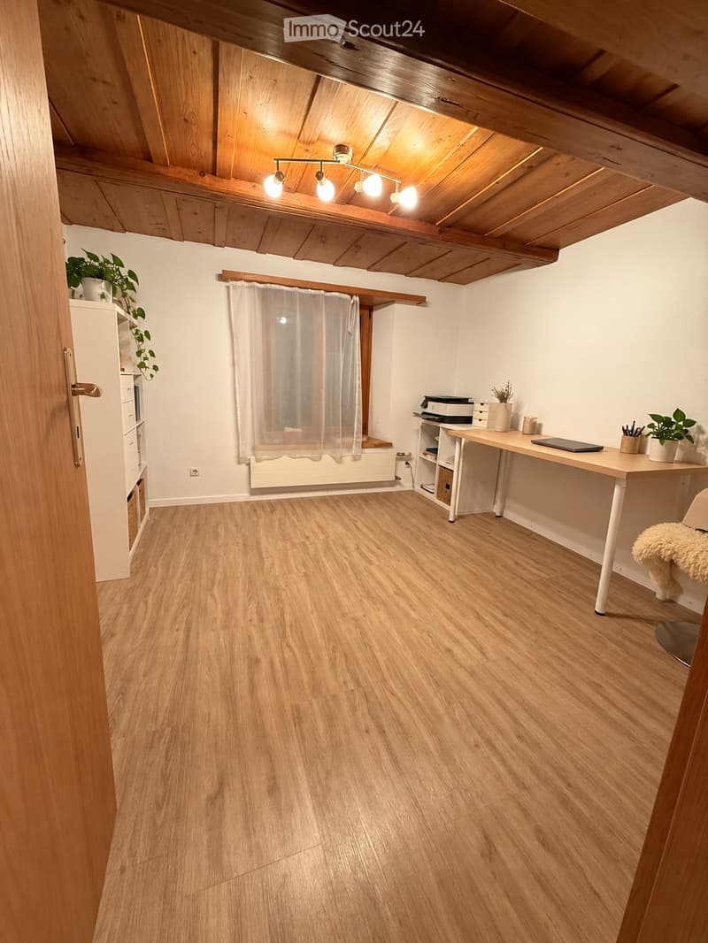 Bright, wooden-floored room with a slanted wooden ceiling and exposed beams. Features a desk with a laptop, chair, small shelf, and potted plants. A window with a curtain and a radiator below it.