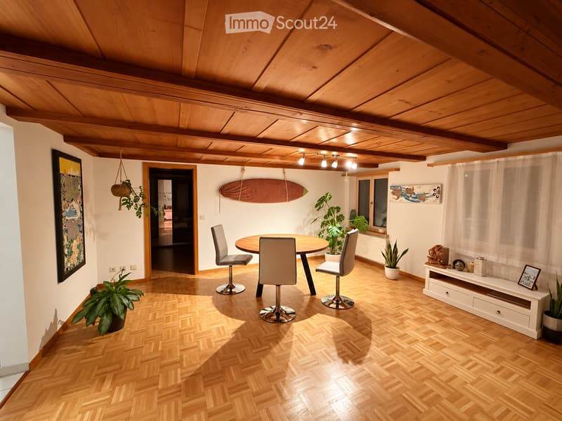 A modern dining area with a round wooden table, four chairs, potted plants, a shelf with decorative items, and a surfboard on the wall.