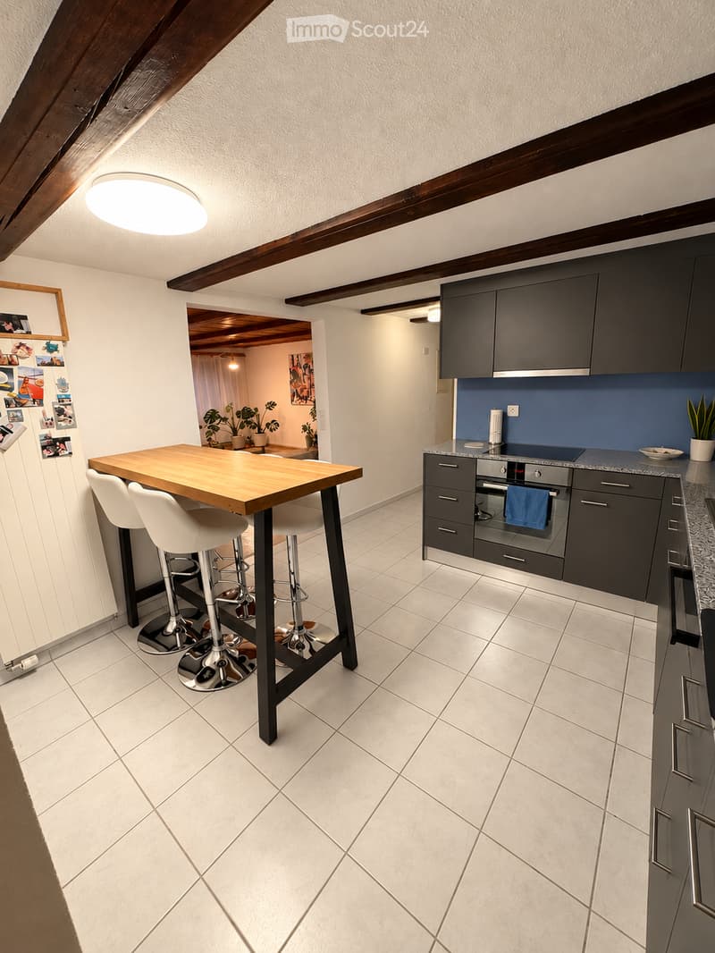 Modern kitchen with dark grey cabinets, a central wooden island table with white stools, and a built-in oven. The space features white tiled flooring and exposed wooden beams on the ceiling.