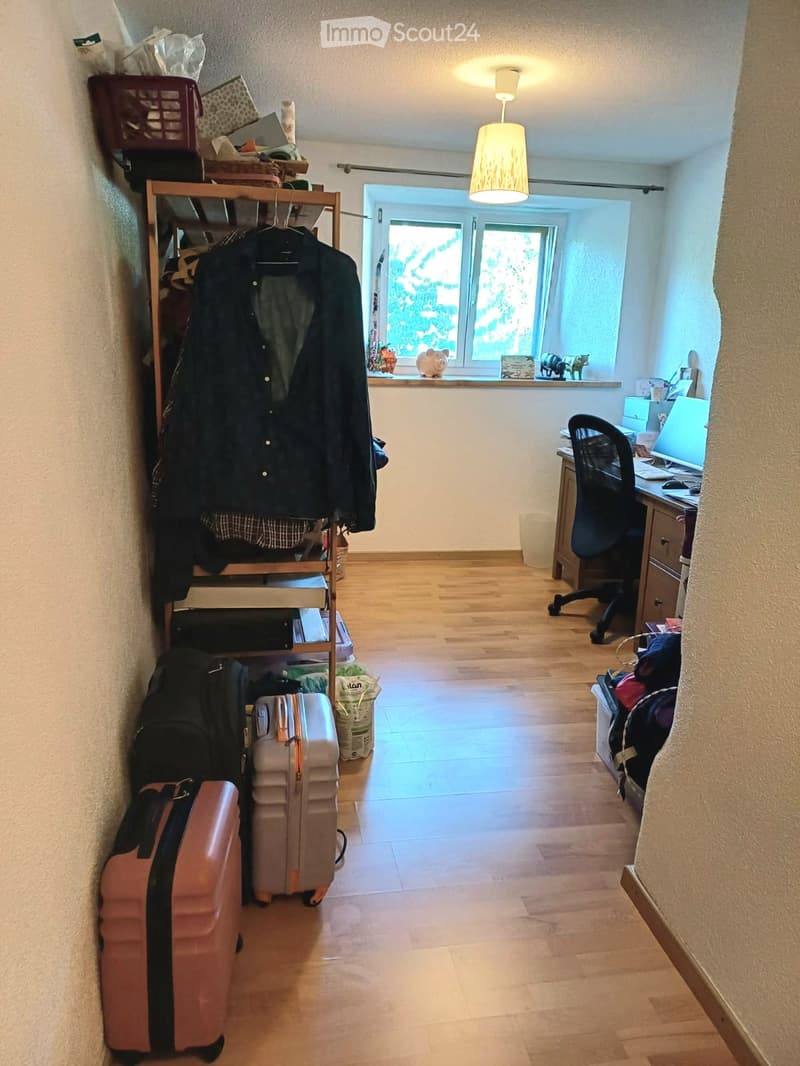 A furnished room with wooden flooring, a desk with a chair and laptop, a clothing rack with jackets, and two suitcases placed near the entrance.