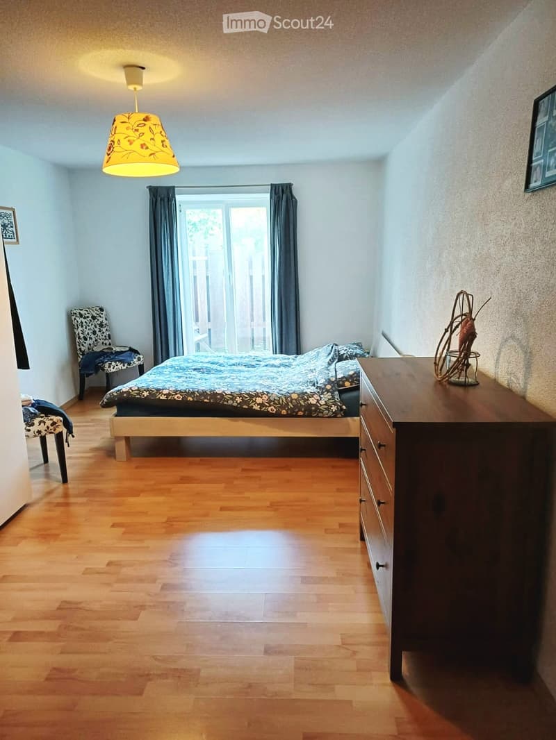 Cozy bedroom with wooden flooring, a bed with patterned bedding, a wooden dresser, a patterned chair, and a hanging lamp with a yellow shade.