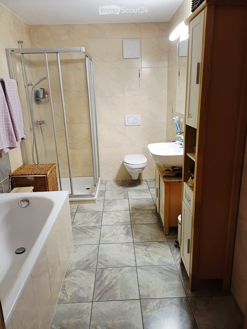 Modern bathroom with a bathtub, toilet, sink, and a glass-enclosed shower. Features a wooden cabinet, wicker basket, and tiled floors and walls.