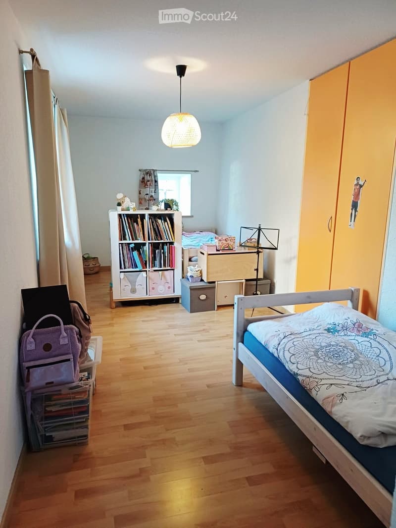 A cozy bedroom with a wooden bed, bookshelf, hanging lamp, and various storage items. The room features light blue walls, wooden flooring, and a window with curtains.