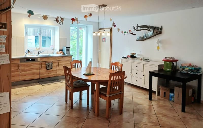Bright kitchen with wooden cabinets, a round wooden dining table surrounded by wooden chairs, a white sideboard, and a black table with books and decorative items. The space features a dishwasher, sink, and a hanging light fixture.
