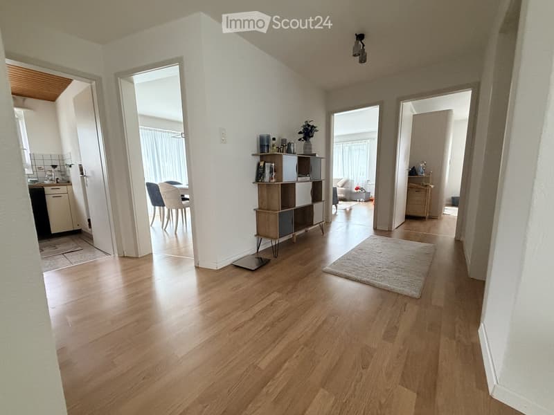 Bright hallway with wooden flooring, leading to multiple rooms including a kitchen, dining area, and living room. Features a small shelf with decorative items and a rug in the center.