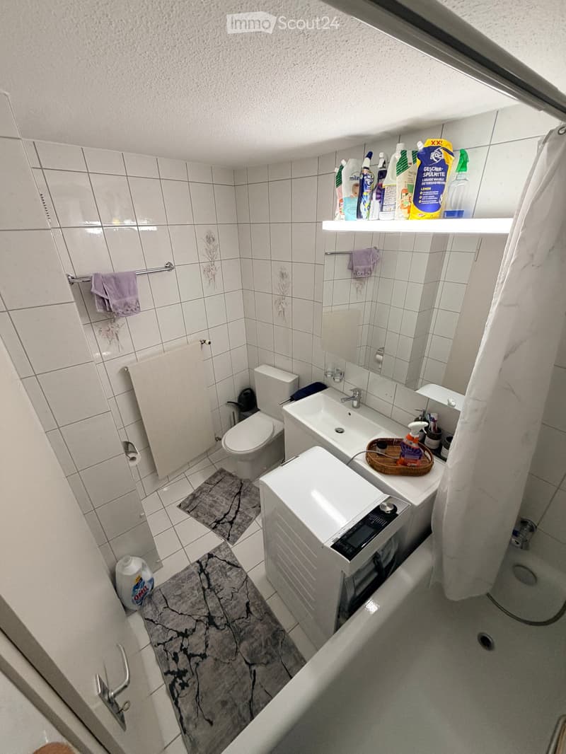 A compact bathroom with white tiled walls and floor, featuring a toilet, sink with mirror, washing machine, shower, and cleaning supplies on shelves.