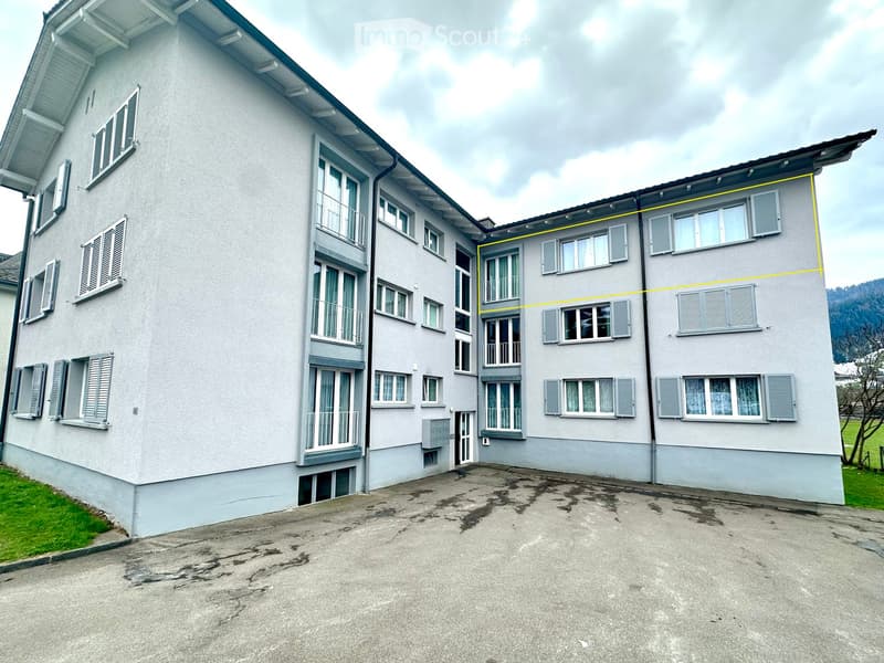 Two-story residential building with light gray facade, multiple windows with shutters, and balconies on the upper floor.