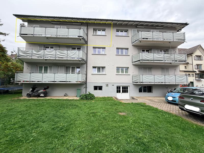 Four-story residential building with multiple balconies, covered with white curtains, and a grassy front yard with parked vehicles.