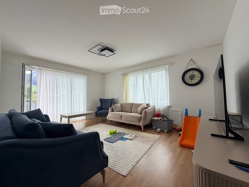 Modern living room with large windows offering a scenic view, featuring a dark blue sectional sofa, a light-colored loveseat, a coffee table, an armchair, a large flat-screen TV, and a decorative clock on the wall.