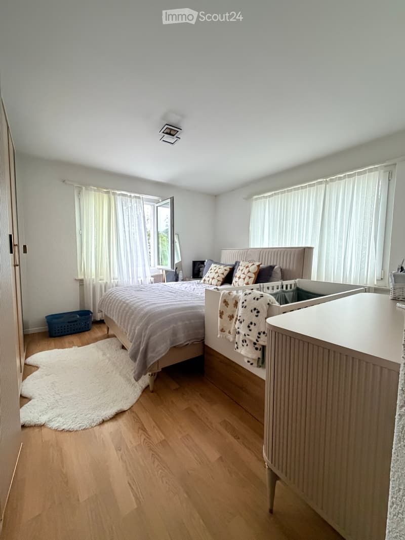 A well-lit bedroom with wooden flooring, a large bed with patterned bedding, a nightstand with a mirror, a baby crib, and a white shag rug near the bed.