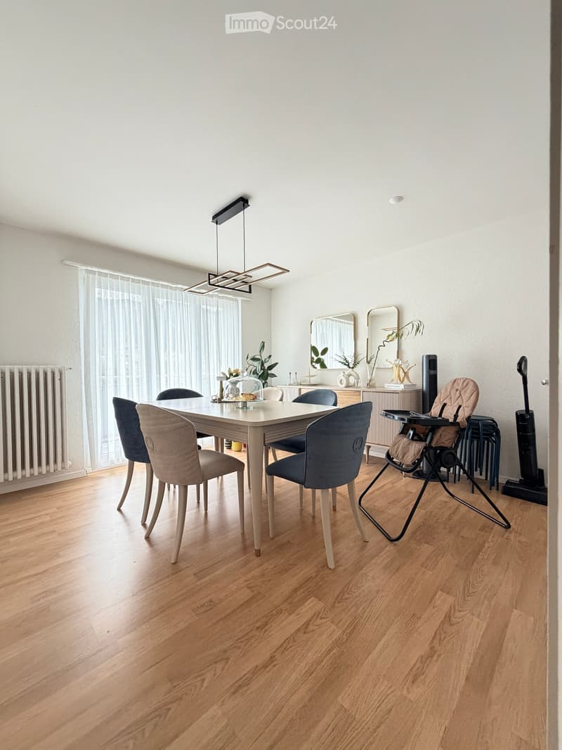 Dining room with a white dining table, four chairs, a baby chair, a mirror, a plant, and a radiator. The room has wooden flooring and large windows with sheer curtains.