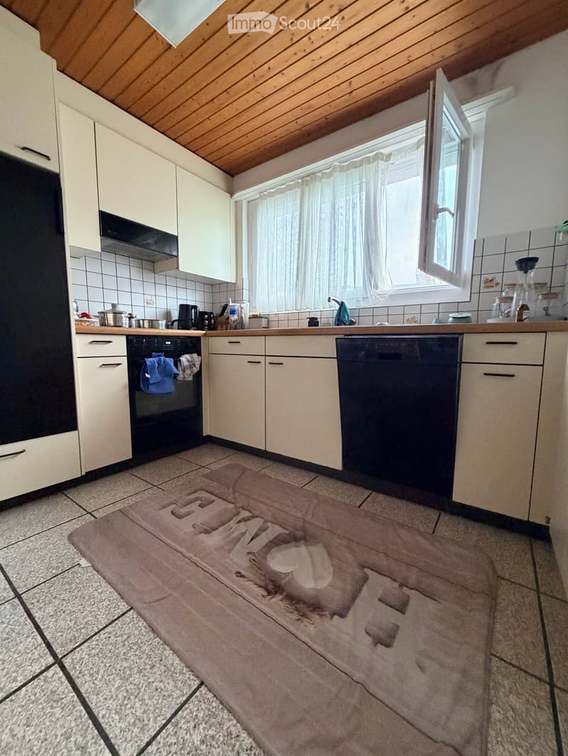 Modern kitchen with white cabinets, stainless steel appliances, tiled backsplash, and a window with a curtain. A large, patterned rug is placed on the tiled floor.
