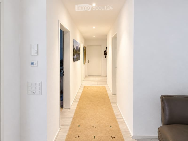 A modern hallway with white walls, tiled floor, and a beige runner rug with small deer and tree designs. There are several wall-mounted light switches and a thermostat on the left wall, and a painting on the right wall. A closed door is visible at the end