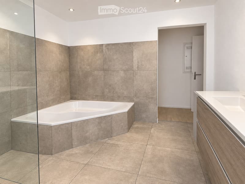 Modern bathroom with a bathtub, sink, and wooden cabinet. The space features tiled walls and floor, with a glass-enclosed shower area.
