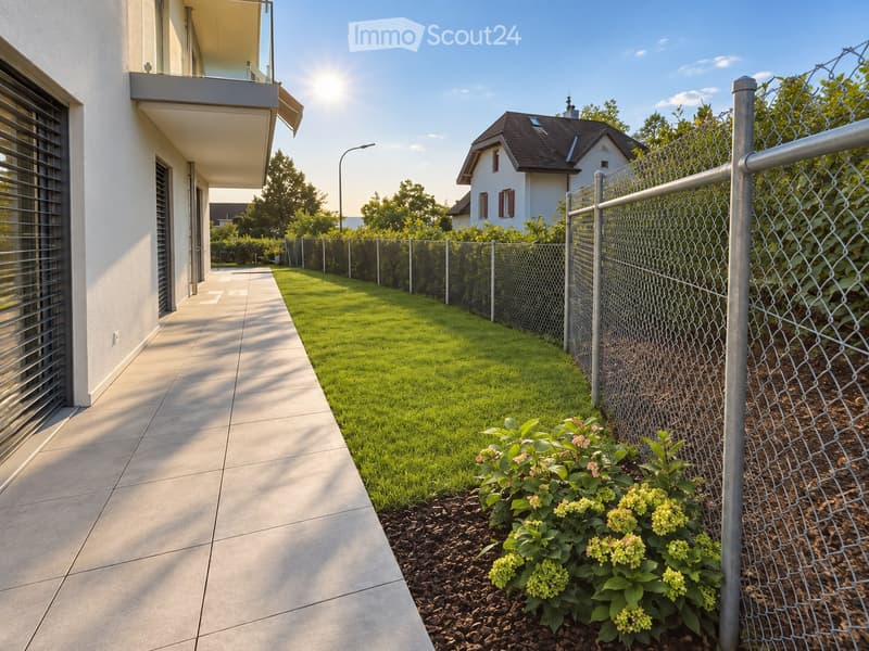 A modern garden area with a well-maintained lawn, a metal fence, and a small flower bed with green and yellow blooms. The pathway is paved with large concrete tiles, and a balcony is visible on the left side.