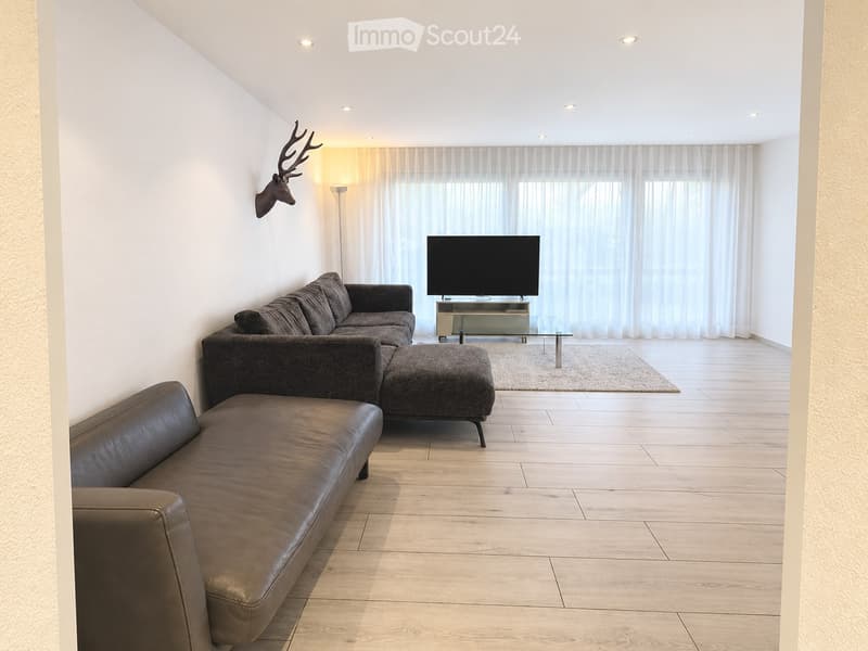 Modern living room with light wooden flooring, large windows with white curtains, a grey sectional sofa, leather sofa, ottoman, TV stand, and a wall-mounted deer head.