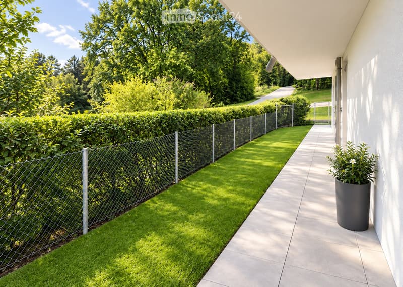 A well-maintained garden area with a neatly trimmed hedge, a metal fence, a paved pathway, and a potted plant near the house.