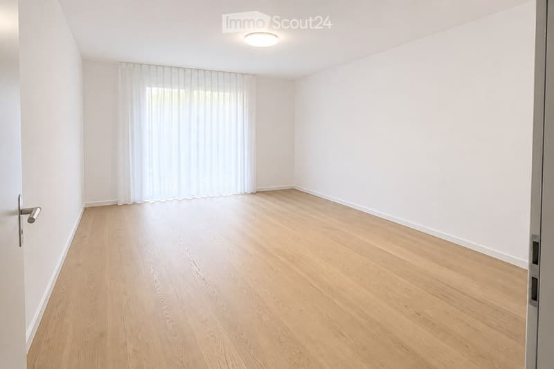 Empty room with light wooden flooring, white walls, and a large window covered by sheer white curtains. A round ceiling light fixture is visible.