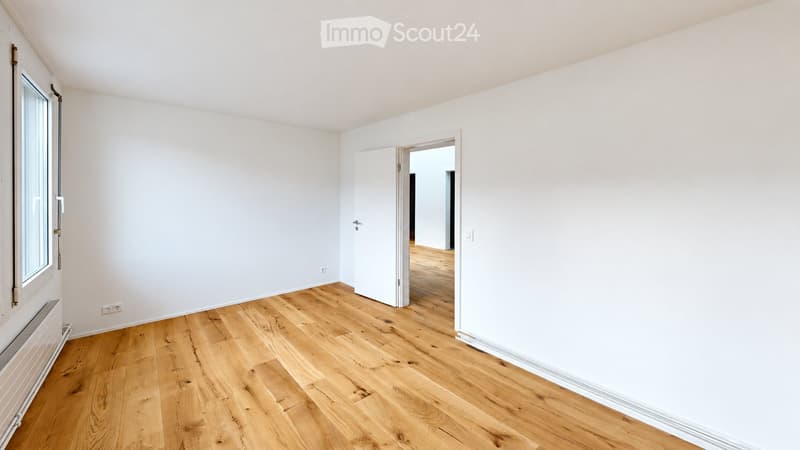 Empty room with light wooden flooring, white walls, and a large window on one side. The room has a door leading to another space.