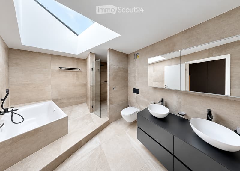 Modern bathroom with a large skylight, beige tiles, a bathtub, a toilet, two sinks, a vanity cabinet, and a mirrored wall.