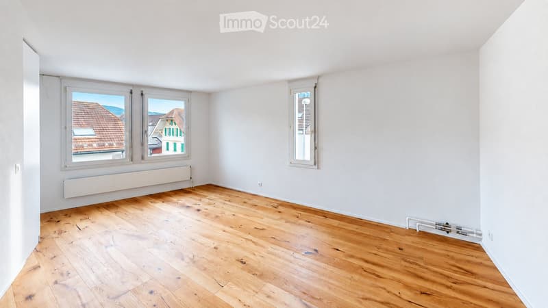 Empty room with wooden flooring, white walls, and large windows offering a view of rooftops and mountains.