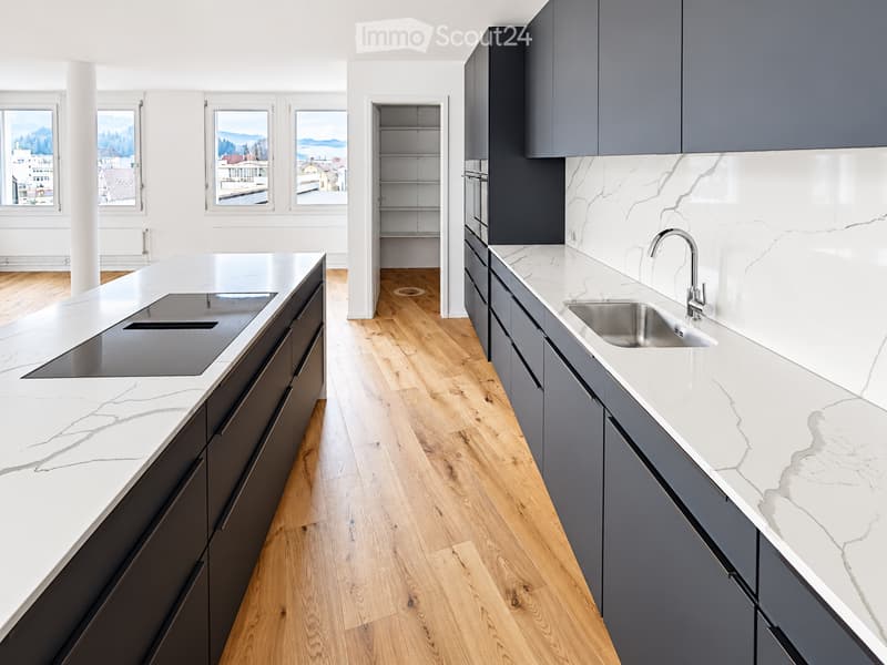Modern kitchen with white marble countertops, dark grey cabinets, stainless steel sink, and induction cooktop. Large windows offer a scenic view.