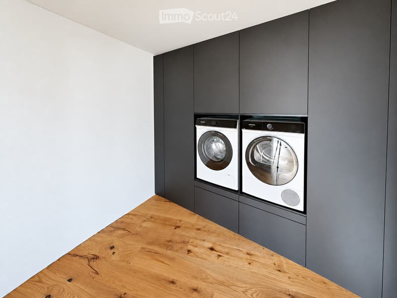 Modern, minimalist room with light wooden flooring and dark grey cabinetry housing a washing machine and dryer.