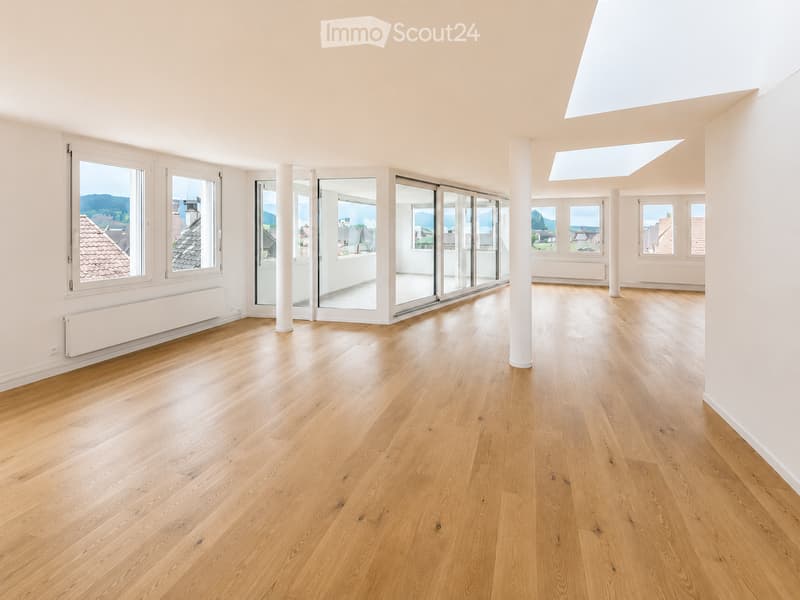 Bright, empty room with light wooden flooring, large windows, and sliding glass doors leading to a balcony. The space features white walls, a skylight, and a radiator on one wall.