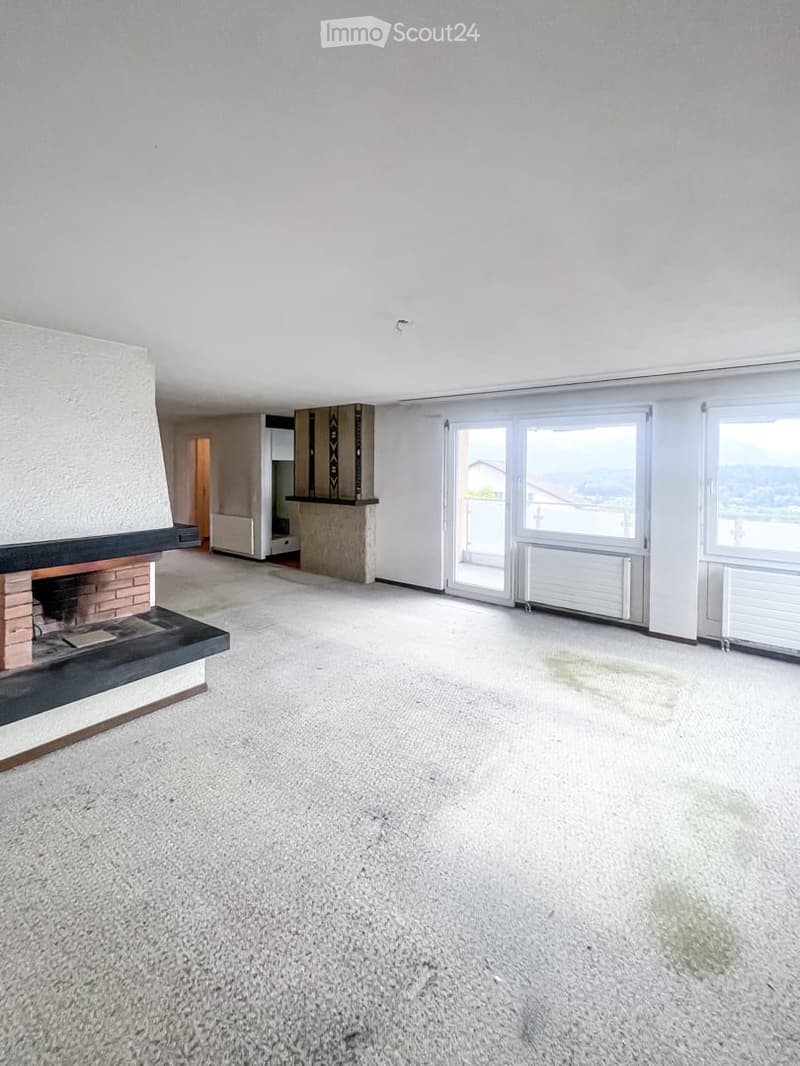 Empty room with a fireplace, large windows offering a scenic view, and a carpeted floor.