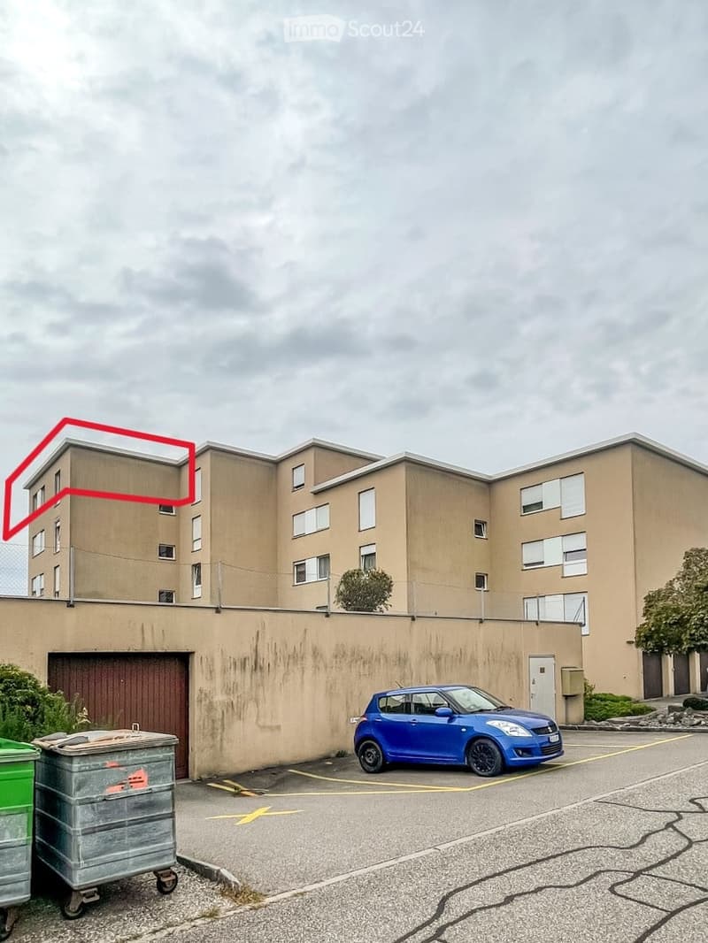 Multi-story residential building with beige facade, multiple windows, and a parking area with a blue car.