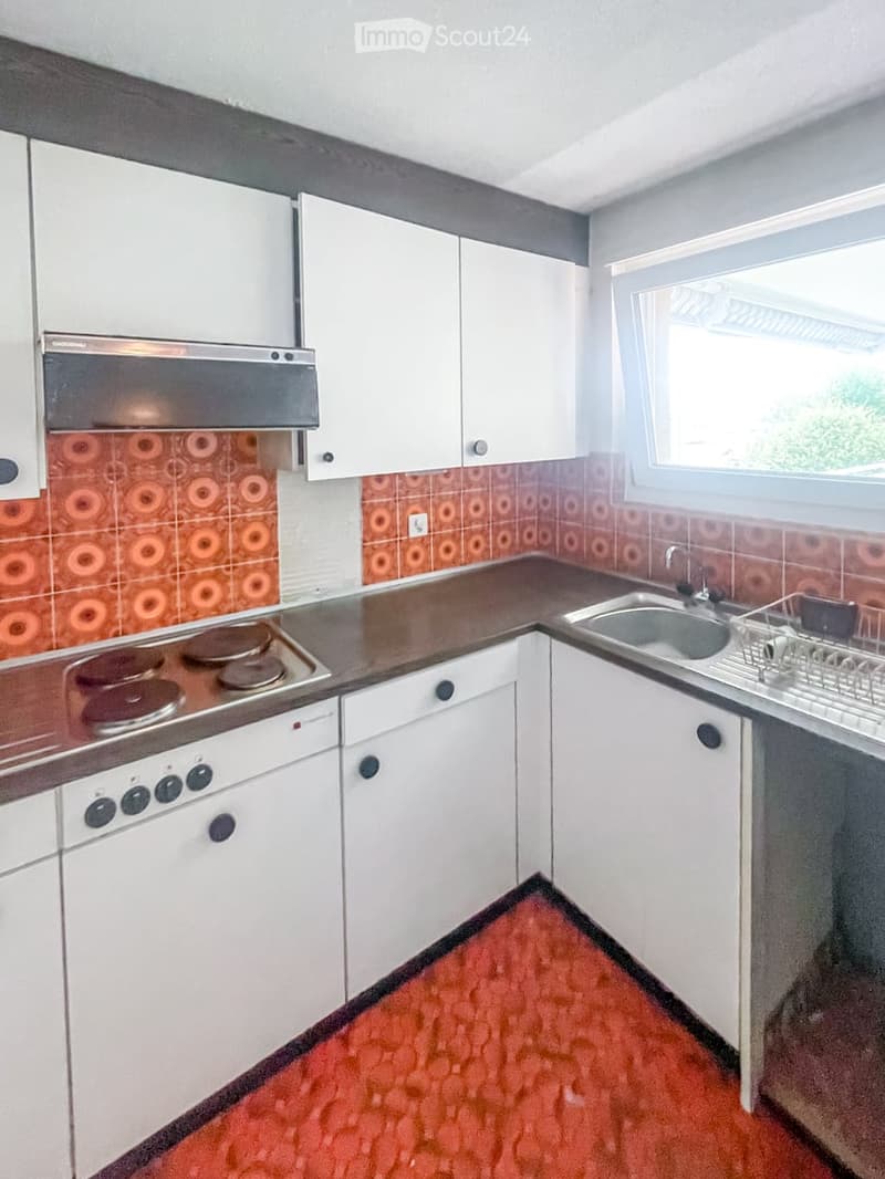 A compact kitchen with white cabinets, a stainless steel stove, a sink, and a dish rack. The backsplash features a retro orange pattern, and the floor is covered in red tiles.