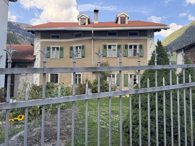 Two-story house with red-tiled roof, green shutters, and ornate metal fence in front.