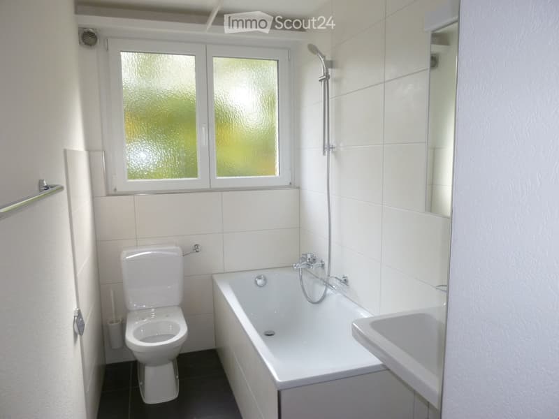 Modern bathroom with white tiles, a bathtub, toilet, sink, and a showerhead. Features a frosted window and a towel rack.
