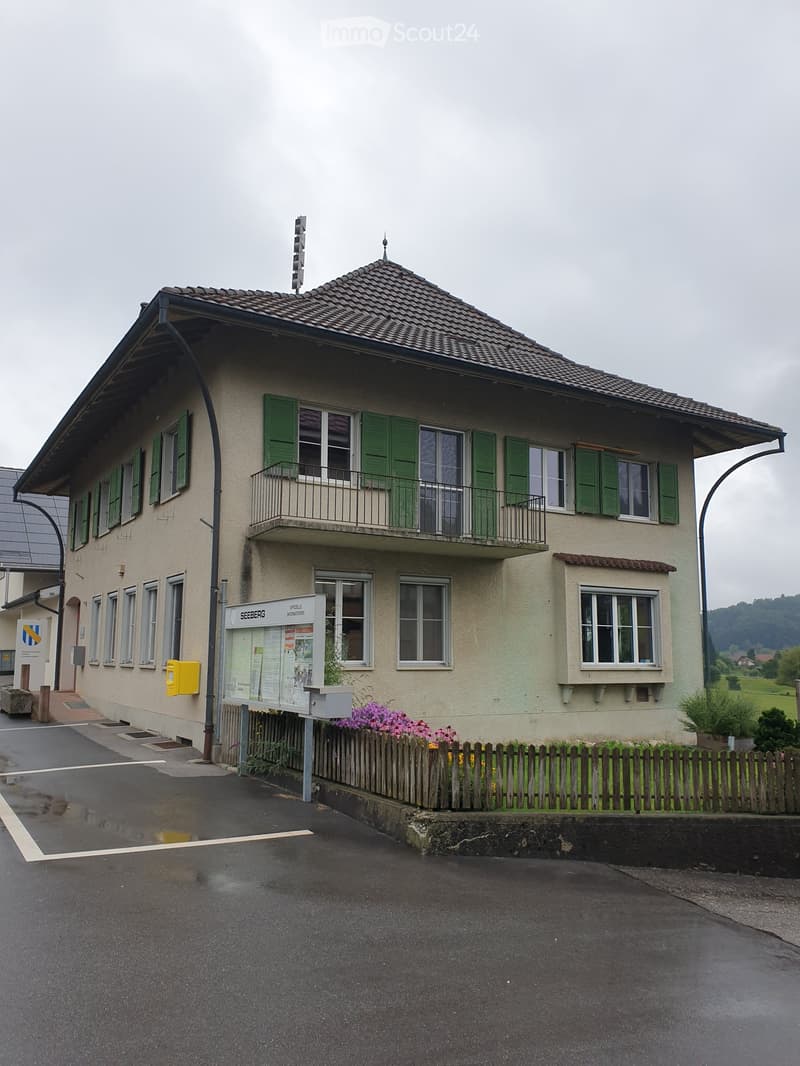 Two-story building with beige walls, green shutters, and a tiled roof. Features a balcony on the upper floor and a small garden area with flowers in front.