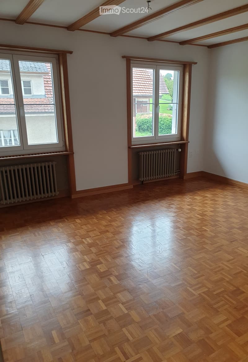 Empty room with wooden herringbone flooring, exposed wooden beams on the ceiling, and large windows with radiators beneath them.