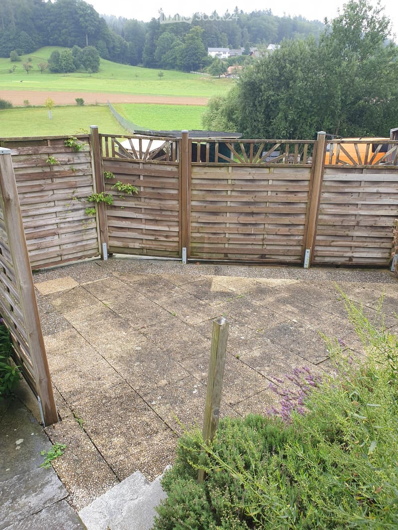 A paved outdoor area with a wooden gate leading to a grassy field, surrounded by trees and shrubs.