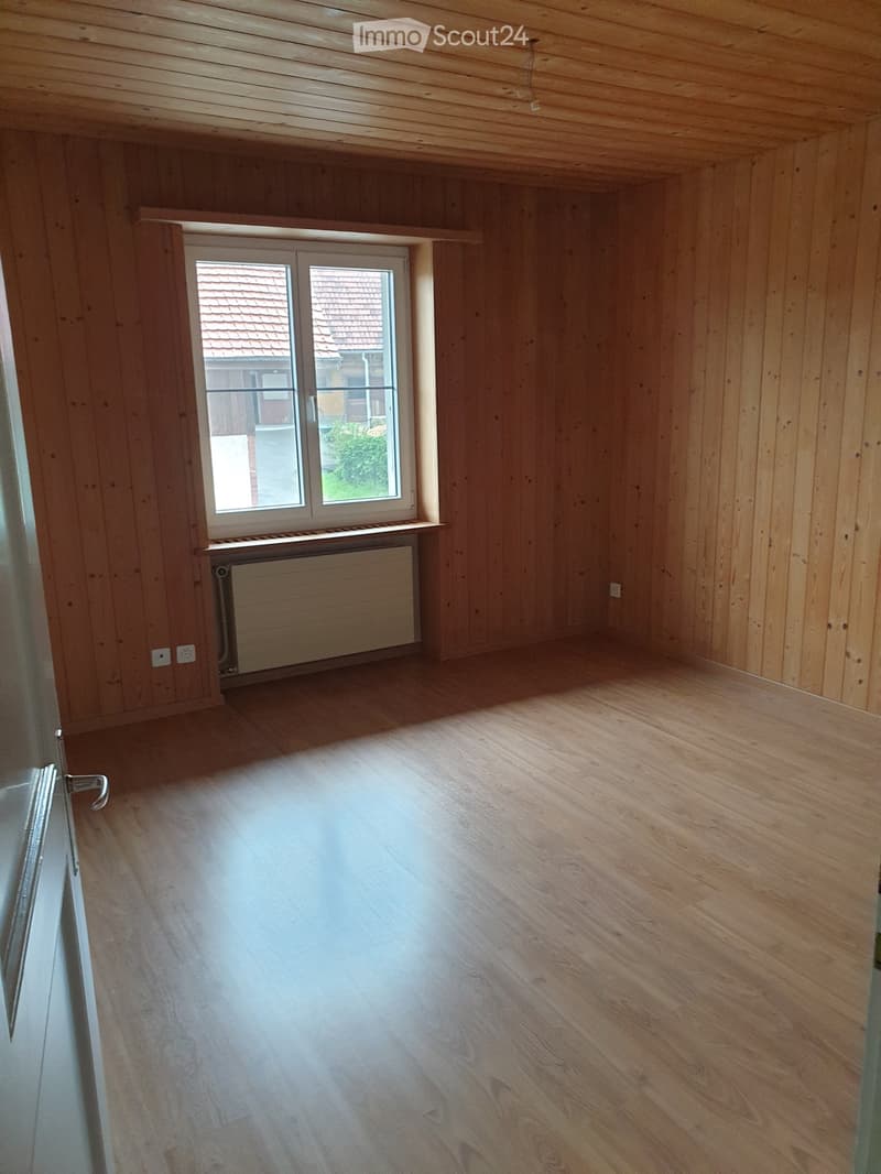 Empty room with wooden walls and floor, large window with view of neighboring house, radiator below window, and visible electrical outlets.