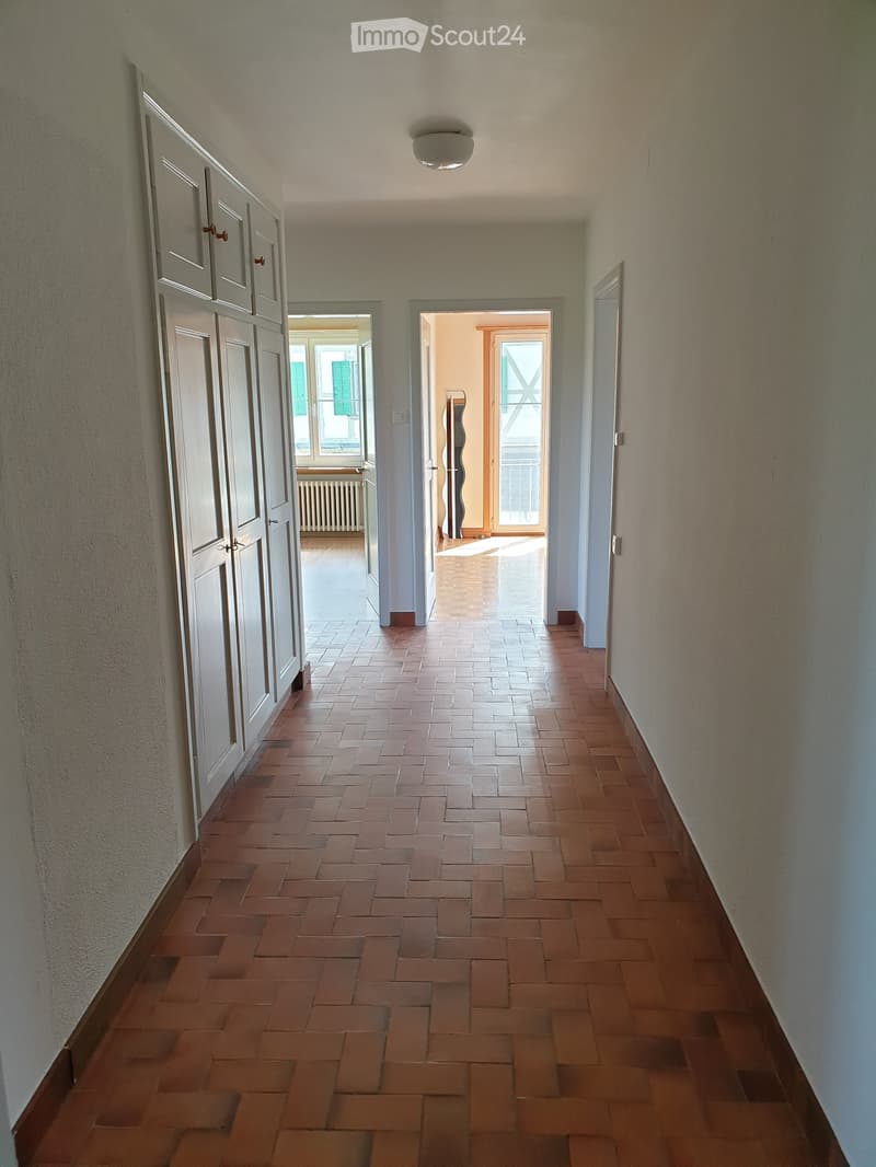 A long, narrow hallway with tiled flooring and white walls, featuring several doors and a ceiling light fixture.