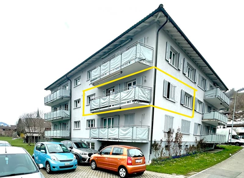 Multi-story residential building with multiple balconies and parked cars in front.