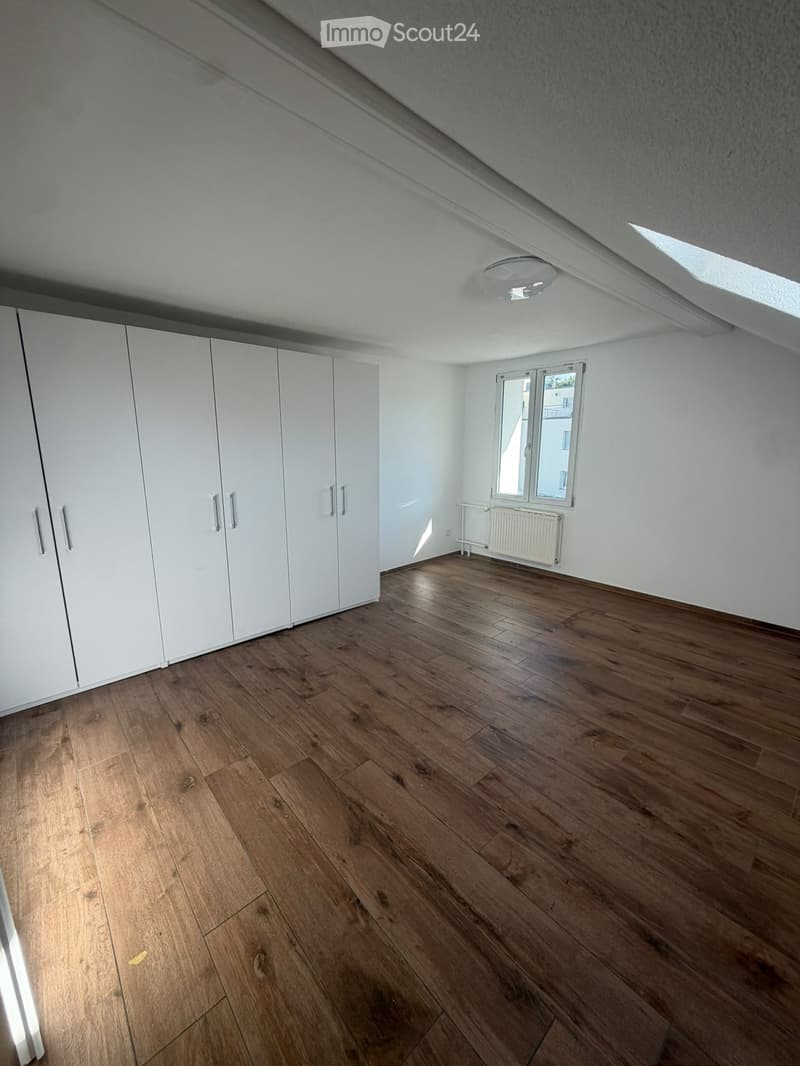 Empty room with wooden flooring, white walls, built-in white wardrobes, a window with a radiator below, and a skylight in the ceiling.
